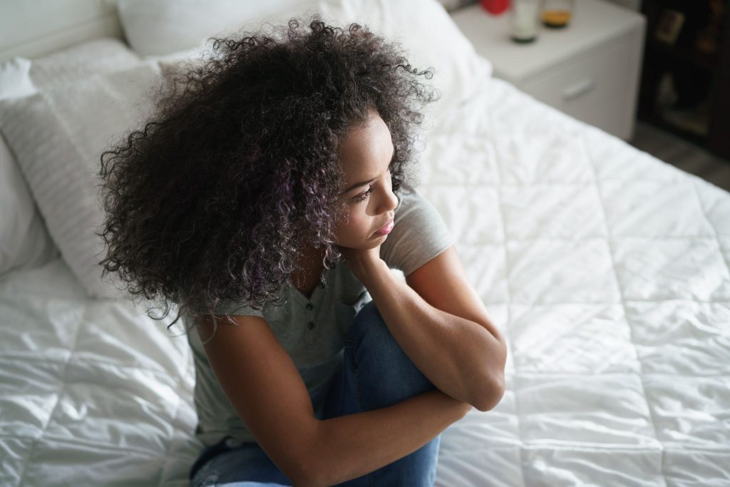 A girl with curly hair sitting on a bed and staring into the distance.