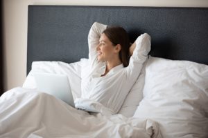 A person sitting up in bed with her arms behind her head and looking out the window